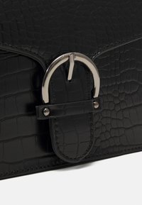 Černá kabelka vyrobená z texturované imitace kůže, s kulatou stříbrnou sponou a prošívanými detaily. Kompaktní obdélníkový tvar.