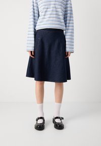 Jeanskjørt med en utsvingt form, mørk blå farge, frontlommer og kontraststing, kombinert med en stripete langermet topp og svarte sko.