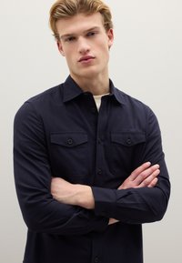 Donkerblauw overhemd van geweven stof met een knoopsluiting aan de voorkant, twee borstzakken en lange mouwen met knoopmanchetten.
