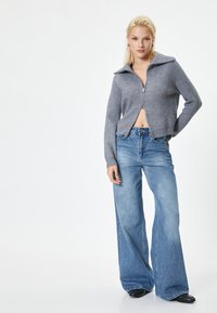 Grå ribbet sweater med krave og lynlås, parret med lyseblå brede bukser og sorte sko. Glat tekstur, afslappet stil.