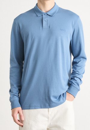 Man draagt een langarmige, blauwe polo met een twee-knops kraag en het "BOSS" logo op de borst, gecombineerd met lichte broek.