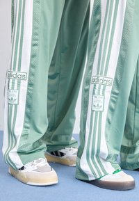 Calças de treino em malha verde com painéis brancos, apresentando a marca Adidas e sapatilhas em contraste, exibindo várias texturas e estilos.