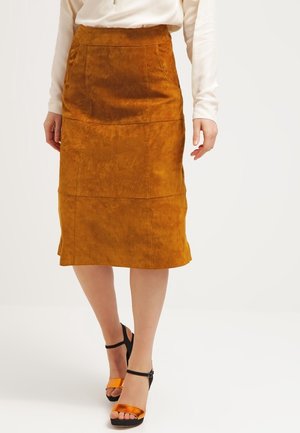 Kvinde iført en rustrød ruskindnederdel til knæene med sømme og forlommer, kombineret med hvid top og sorte og orange sandaler med hæl.