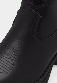 Κοντινή λήψη ενός λείας μαύρης δερμάτινης μπότας με ελαστικό πλαϊνό πάνελ και διακοσμητικό ράψιμο στην περιοχή των δαχτύλων.