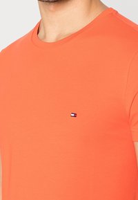 T-shirt di cotone arancione con scollatura rotonda e un piccolo logo ricamato in rosso, bianco e blu sul lato sinistro del petto.