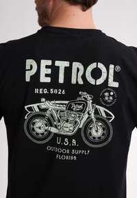 Črna majica s kratkimi rokavi, ki prikazuje belo grafiko motocikla ter besedilo "PETROL", "U.S.A." in "OUTDOOR SUPPLY FLORIDA." Zasnova je obrabljena.