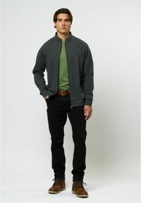 Chaqueta de cremallera gris oscuro sobre una camisa verde, combinada con pantalones negros y botas marrones. Tejido suave, diseño casual y estilo ajustado.
