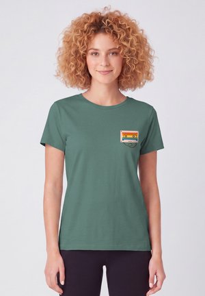 Femme aux cheveux bouclés portant un t-shirt vert avec un petit motif coloré représentant une cassette sur la poitrine, debout devant un fond uni.