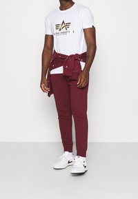 Vit t-shirt med guldfärgad logga, röda mjukisbyxor och vita sneakers. Sweatshirt knuten runt midjan med ett ljust stänkmönster.