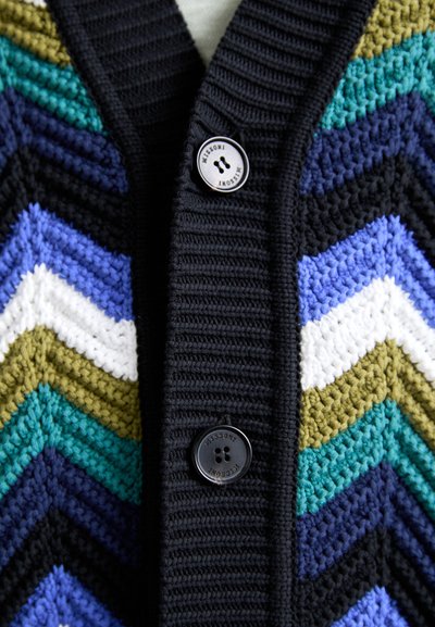 Cardigan croșetat cu un model în zig-zag în negru, verde, violet, albastru și alb, cu o textură în cută și două butoane negre.