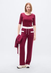 Σετ αθλητικών ρούχων μπορντό με εφαρμοστή μπλούζα και φαρδιά παντελόνια. Η μπλούζα έχει μανίκια τρία τέταρτα, ενώ το παντελόνι διαθέτει κορδόνι στη μέση και διπλές πλαϊνές τσέπες.