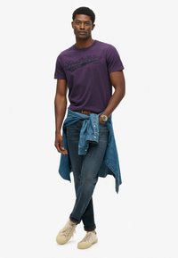 Fioletowa koszulka z krótkim rękawem z czarnym logo, w zestawieniu z niebieskimi dżinsami i jasnoniebieską koszulą dżinsową związaną w talii. Beżowe trampki dopełniają całość.