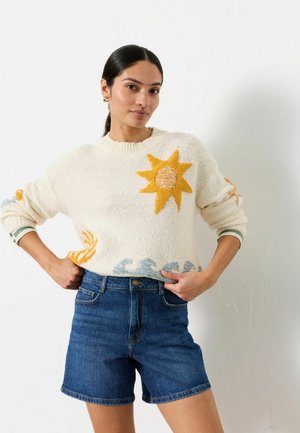 Woman with dark hair in a ponytail wears a white sweater with sun and wave patterns and high-waisted blue denim shorts, posing with hands on hips.