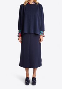 Frau in einem übergroßen marineblauen Oberteil und einem Midikleid mit bunten Blumenmuster an den Ärmeln und glänzenden navyblauen Plateauslippern.
