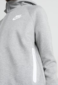 Grå zip-up hoodie gjord av mjukt tyg, med en vit Nike-logga, en framficka med vit dragkedja och en huva.