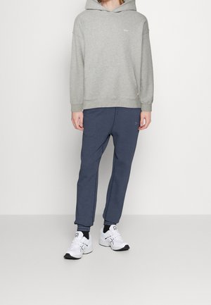 Grå hoodie med dragsko och logotyp, kombinerad med marinblåa joggers och vita sneakers. Outfiten har en avslappnad passform och mjuk textur.