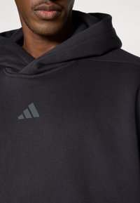 Μαύρο φούτερ με υφασμάτινη υφή, που διαθέτει ένα εξέχον γκρι τριγωνικό λογότυπο στο στήθος και μια στρωματοποιημένη κουκούλα για επιπλέον βάθος.