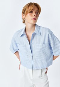 Camicia a maniche corte in blu chiaro con righe orizzontali bianche, dotata di due tasche frontali e maniche larghe.
