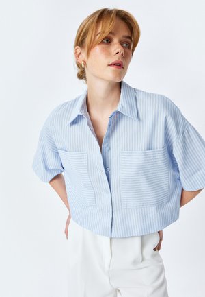 Cropped button-up shirt in light blue with vertical white stripes, featuring two front pockets and short, wide sleeves.
