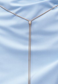 Λεπτό χρυσό κολιέ lariat με μικρό κυκλικό κούμπωμα σε φόντο λείας ανοιχτό μπλε υφής.