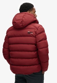 Veste doudoune rouge avec capuche, présentant un design matelassé et des coutures visibles. Marque "SUPERDRYJP" dans le dos en couleurs contrastées.