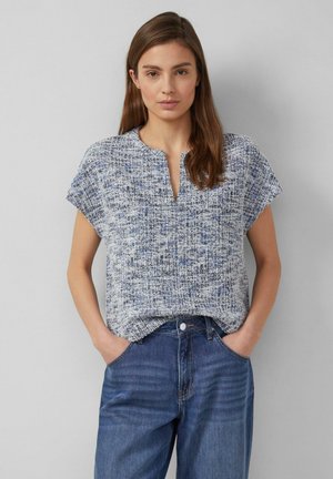 Femme portant un haut bleu et blanc texturé à manches courtes avec une encolure fendue et un jean bleu, debout les mains dans les poches.