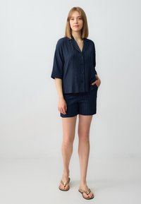 Top blu navy a maniche corte con bottoni e colletto, abbinato a pantaloni corti. Presenta righe verticali e una vestibilità comoda. Include tasche laterali.