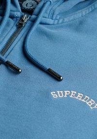 Felpa con zip blu realizzata in tessuto morbido, con logo "SUPERDRY" curvato in rosa chiaro e zip nera con testo in rilievo.