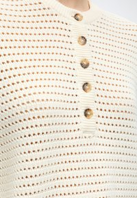 Μπλούζα μπεζ χρώματος με πλεκτό ανοιχτό σχέδιο και πέντε κουμπιά από χελώνα μπροστά κοντά στον γιακά.