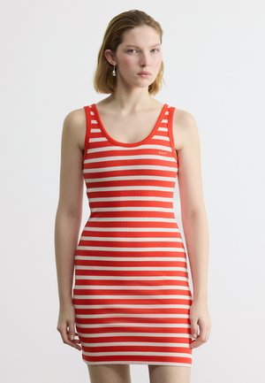 Νεαρή γυναίκα που φοράει κόκκινο και λευκό οριζόντιο ριγέ φόρεμα χωρίς μανίκια, που στέκεται μπροστά σε ένα απλό φωτεινό φόντο.