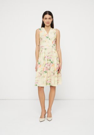 Frau mit langen dunklen Haaren, die ein ärmelloses, pastellfarbenes, knielanges Kleid mit Blumenmuster und beige High Heels trägt, steht vor einem weißen Hintergrund.