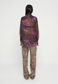 Camicia trasparente viola con motivo a foglie di palma, indossata sopra pantaloni a gamba larga beige con stampa leopardata. Vestibilità ampia, maniche lunghe e colletto.