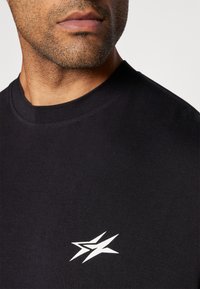Černé bavlněné tričko s kulatým výstřihem a bílým grafickým logem na levé straně hrudi. Hladká textura, minimalistický design.