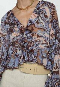 Blouse à imprimé floral avec des détails à volants en marron, bleu et rose sur un tissu transparent ; portée avec une ceinture beige soulignant la taille.
