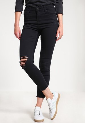 Mujer con jeans ajustados negros rotos y zapatillas blancas, de pie con las piernas cruzadas frente a un fondo liso.