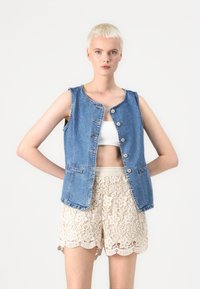 Denimvest med rund halsutskjæring og frontknapper, med to sidelommer. Kombinert med kremfargede blondershorts med bølgede kanter.