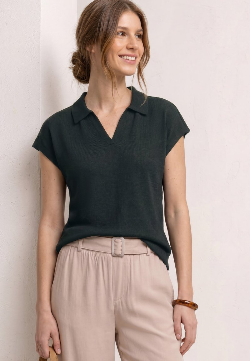 Femme aux cheveux bruns en chignon, portant un haut noir à manches courtes avec col et un pantalon beige ceinturé, souriante et debout contre un mur clair.