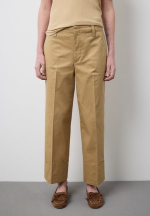 Person trägt beige Hosen mit hoher Taille und weitem Bein, die am Saum umgeschlagen sind, kombiniert mit braunen Mokassin-Schuhen und einem ärmellosen beigen Oberteil.