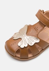 Brązowy skórzany sandał z motylkowym wzorem w kolorze metalicznego złota, z wycięciami po bokach oraz zapięciem na rzep.