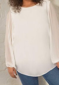 White blouse with pleated long sleeves, soft texture, and a rounded hem, paired with blue denim jeans.