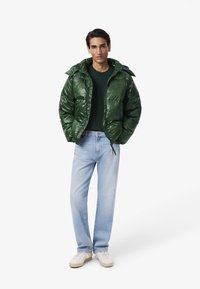 Giacca a vento verde lucido con cappuccio, indossata sopra una maglietta scura. Abbinata a jeans azzurri chiari e sneakers bianche. Design semplice e contemporaneo.