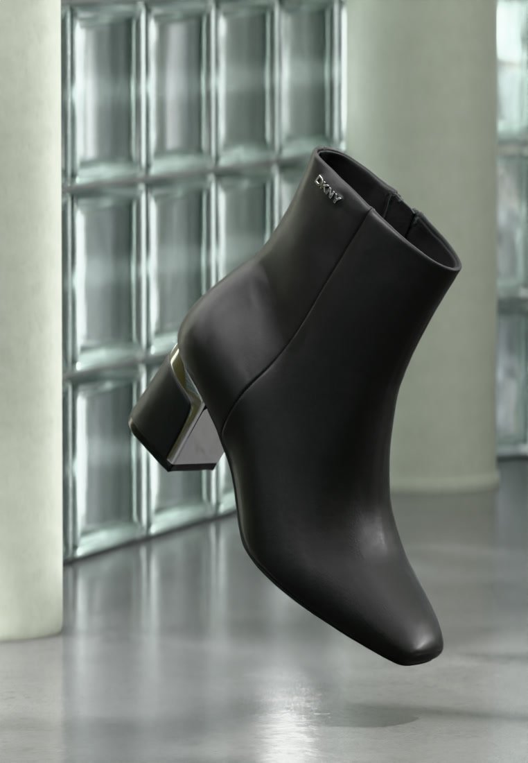 Bottine en cuir noir avec talon bloc et logo DKNY argenté près du haut, posée devant un mur en briques de verre et un sol lisse.