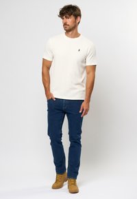 Lys beige kortærmet T-shirt med rund hals og lille logo, parret med mørkeblå slim-fit jeans og brune snørrede støvler.
