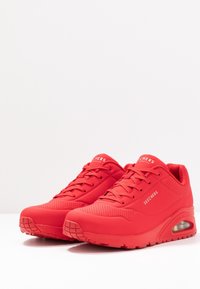 Ett par röda Skechers sportskor med snörning, perforerade detaljer och synliga luftkuddssulor, vinkelrätt på en vit yta.