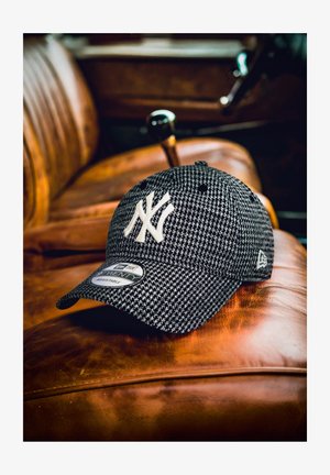 Boné houndstooth preto e branco dos New York Yankees repousado num banco de carro de pele castanha, com a alavanca de mudanças vintage visível.
