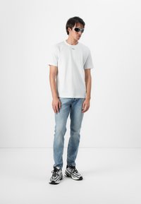 Tricou alb din bumbac cu un mic logo, blugi din denim albastru deschis și pantofi sport alb-negru. Modelul poartă ochelari de soare gri.