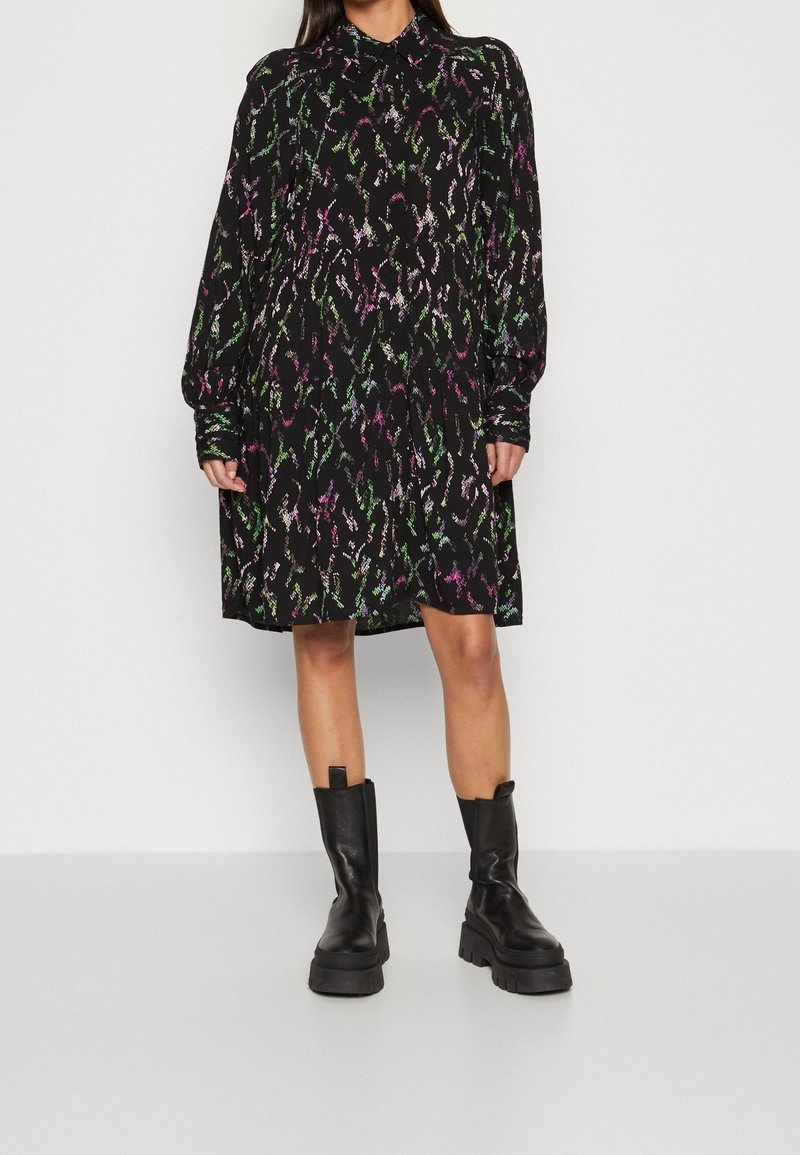 Czarna sukienka z kołnierzykiem, długimi rękawami i swobodną sylwetką. Posiada kolorowe abstrakcyjne wzory w zieleni, fioletach i różu. Stylizowana z masywnymi czarnymi botkami.