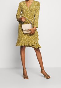 Kvinna i en senapsgul blommig svepklänning med volangkant, håller en beige texturerad handväska och bär olivgröna remsandaler.