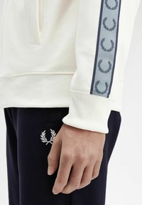Sudadera blanca con puños acanalados, que presenta un panel lateral azul decorado con diseños de laurel. Combinada con pantalones azul marino y detalles de logo.