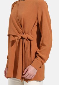 T-shirt à manches longues en tissu couleur rouille avec un devant drapé et un détail noué à la taille. Texture douce et coupe décontractée avec un col rond.